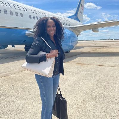 She is taking a picture in front of the president's plane wearing a jacket while carrying a handbag.